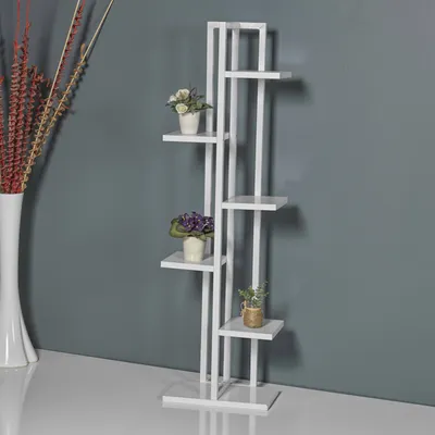 A white plant stand with several shelves is shown against a gray wall. There are three small potted plants on the stand. To the left, a tall white vase with dried flowers is visible.