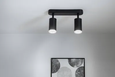 A black ceiling light fixture with two adjustable spotlights is mounted on a white ceiling. Below the light fixture, a framed piece of abstract art with circular patterns is visible.