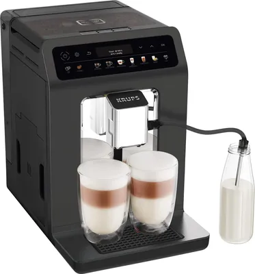 A black KRUPS Evidence One automatic coffee machine is shown with two glasses of latte macchiato and a milk carafe connected by a tube.