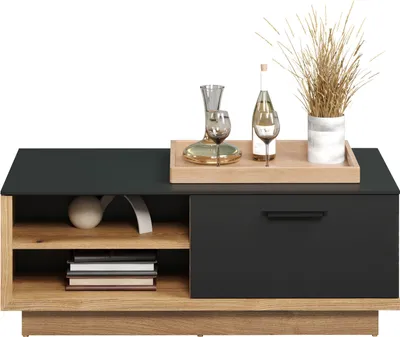 A modern coffee table with a dark top and wooden base, decorated with a decanter, wine glasses, a bottle, and dried grass in a vase.