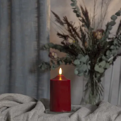 A dark red LED candle with a flickering flame sits on a draped gray fabric, with a blurred background of a curtain and a bouquet of dried flowers.