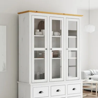 A white cabinet with glass doors and a wooden top, displaying some kitchenware inside. The cabinet is placed in a room with a light gray wall and a hint of a sofa and a small table in the background.