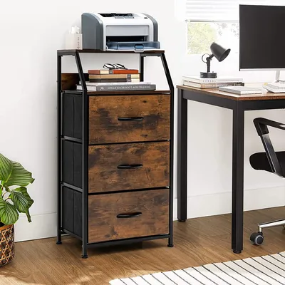 A storage cabinet with fabric drawers and open shelves is placed next to a desk with a computer and a lamp.