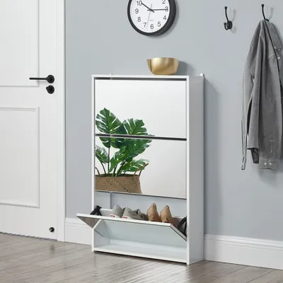 A white shoe cabinet with mirrored doors is shown in a hallway next to a white door. A plant is visible inside the cabinet, and shoes are stored in the open bottom compartment. A clock and coat hooks are on the wall above.