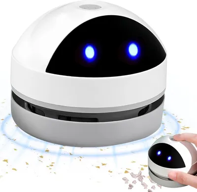 A white and gray robotic-shaped mini vacuum cleaner is shown on a white surface with small crumbs around it. A hand is holding a smaller version of the vacuum cleaner.