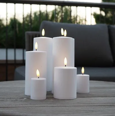 Several white LED candles of varying sizes are arranged on a wooden table outdoors, with a blurred background of outdoor furniture and greenery.