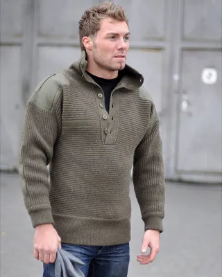 A man wearing an olive green sweater and blue jeans stands outside.
