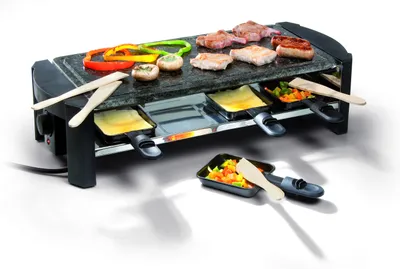 A raclette grill with food items on it, including meat, vegetables, and cheese.