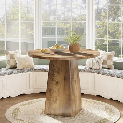 A round wooden dining table with a pedestal base is set in a sunlit bay window area with built-in benches and cushions.