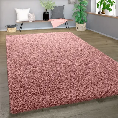 A shaggy pink rug is laid out on a wooden floor in a living room setting. A bench with pillows and a plant are visible in the background.