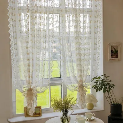 A window with sheer curtains, a table with cups and a vase of flowers, and a plant.