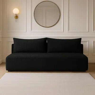 A black sofa with two large pillows, placed in a room with white paneled walls and a round gold mirror.