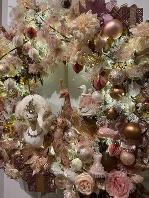 A close-up view of a festive Christmas wreath decorated with various ornaments, including glass baubles, artificial flowers, and decorative birds, all in shades of pink and gold.