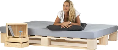 A woman is lying on a bed frame made of wooden pallets, with a pillow and a small bedside table made of wooden crates.