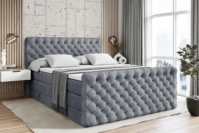 A tufted grey boxspring bed with two white pillows and a grey throw blanket is shown in a bedroom setting.