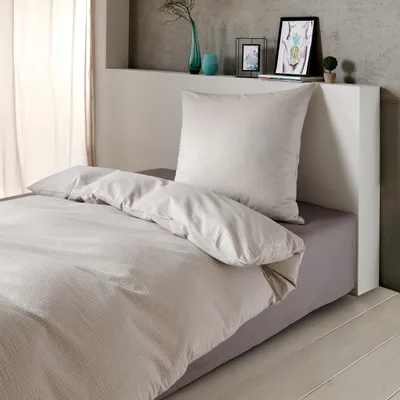 A bed with silver-grey seersucker bedding, including a duvet and pillow, set against a minimalist bedroom background with a shelf holding decorative items.