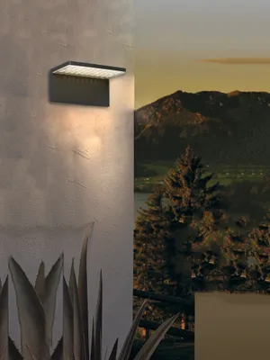 A modern outdoor wall light is mounted on a textured wall, casting a warm glow downwards. The background transitions to a scenic landscape with mountains, a lake, and trees, suggesting the product's intended outdoor use.