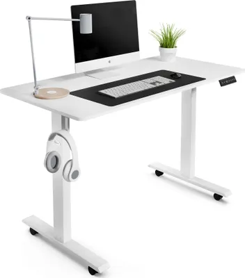 A white adjustable standing desk with a computer, keyboard, mouse, lamp, and headphones.