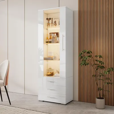 A white high-gloss display cabinet with glass doors and drawers, illuminated from within, stands in a modern room with a slatted wood accent wall and a plant.