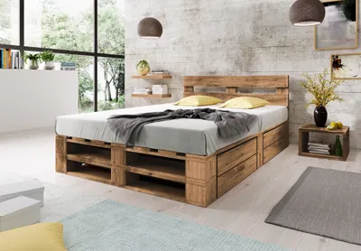 A bedroom scene featuring a wooden pallet bed with storage drawers, set against a textured wall and large windows.
