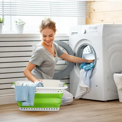 A woman is doing laundry in a laundry room, putting clothes into a washing machine. A green and white laundry basket is in the foreground.