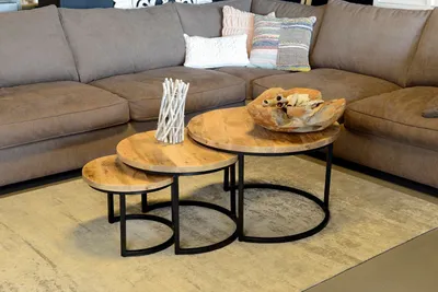 A set of three nesting coffee tables with wooden tops and black metal legs, placed in a living room setting next to a large sectional sofa.