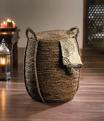 A woven basket with a lid and a decorative cloth draped over it, placed on a wooden floor next to a lantern and a table with candles.