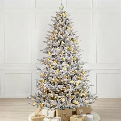 A flocked artificial Christmas tree with warm white LED lights and presents at its base, set against a white paneled wall.