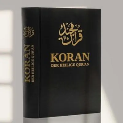 A black book with gold lettering, likely a religious text, is shown against a light gray background with soft shadows.