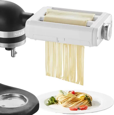 A black KitchenAid mixer attachment is shown cutting pasta dough into fettuccine noodles, with a plate of finished pasta in the foreground.