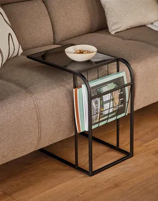 A black metal side table with a glass top and magazine rack is placed next to a brown sofa.