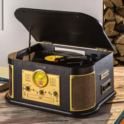 A Technaxx TX-103 multifunctional record player is shown on a wooden surface, with a stack of firewood in the background.