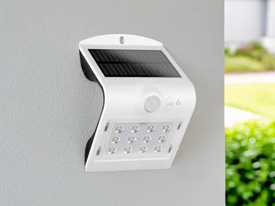 A white solar LED butterfly light with a motion sensor is mounted on a textured gray wall. The background shows a blurred green garden and a white pillar.