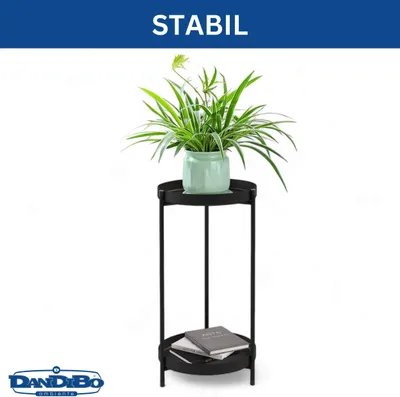 A black metal plant stand with two tiers, holding a potted green plant on the top tier and two books on the bottom tier. The background is white.