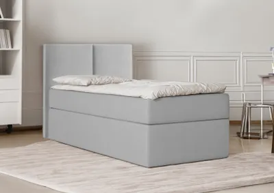 A light gray bed with a headboard is shown in a room with a white wall and a rug.