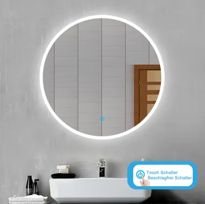 A round LED mirror is mounted on a wall above a sink in a modern bathroom. The mirror has a soft white light around its edge. A small blue dot is visible on the mirror's surface, likely indicating a touch control. Below the mirror is a white sink with a fa