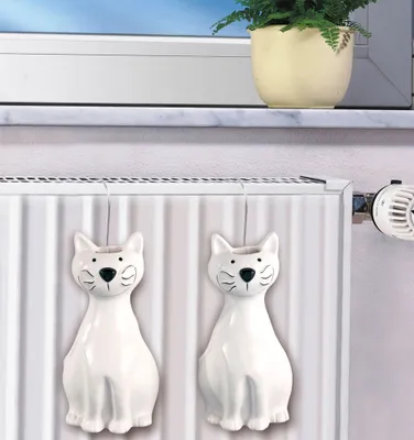 Two white ceramic cat-shaped humidifiers hang on a radiator, with a potted plant visible on a windowsill in the background.