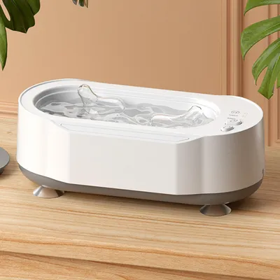 A white and gray ultrasonic cleaner filled with water sits on a wooden surface, with a green leaf visible in the background.