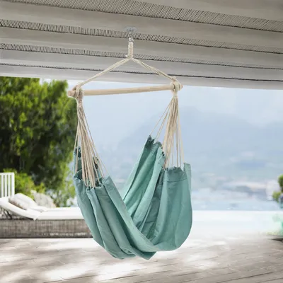 A teal hammock chair is suspended from a wooden beam on a porch, with a blurred background of trees, water, and mountains.