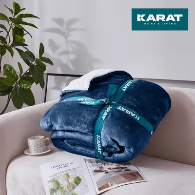 A folded dark blue blanket with a white sherpa lining is placed on a sofa next to a plant and a magazine.