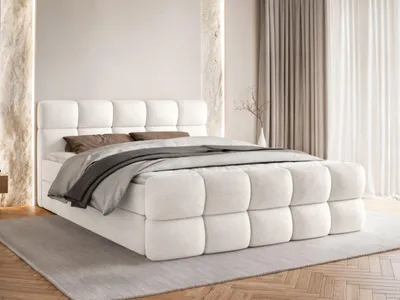 A white boxspring bed with a tufted headboard and footboard, dressed with bedding, in a room with neutral-toned curtains and decorative wall elements.