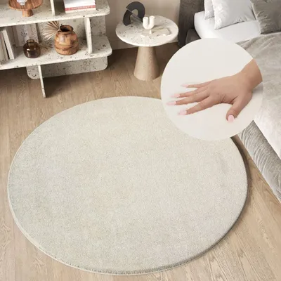 A round, cream-colored rug is shown in a bedroom setting, with a hand gently pressing into its soft surface.