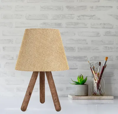 A table lamp with a beige lampshade and wooden legs is placed on a white surface in front of a white brick wall. There are also some paintbrushes in a jar and a small potted plant.