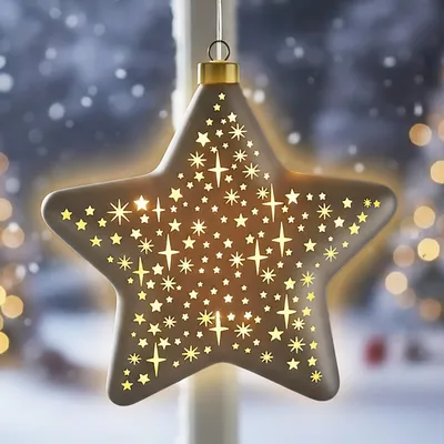 A star-shaped Christmas ornament with cut-out stars, illuminated from within, hanging in a window with a snowy outdoor scene and bokeh lights in the background.