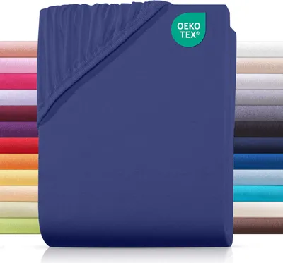 A folded navy blue fitted sheet is shown, with a stack of other colored sheets behind it on the left and a stack of gray-toned sheets on the right. A green circular logo with white text is in the upper right corner.
