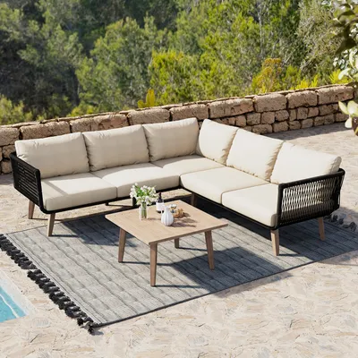An outdoor patio setting with a sectional sofa, coffee table, and rug, with a stone wall and trees in the background.