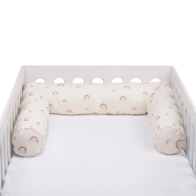 A baby crib with a white mattress and a cream-colored crib bumper with a rainbow pattern.