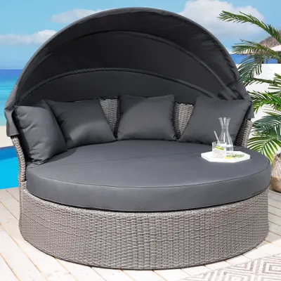 A round outdoor daybed with a canopy and cushions, set on a wooden deck next to a pool and palm trees.