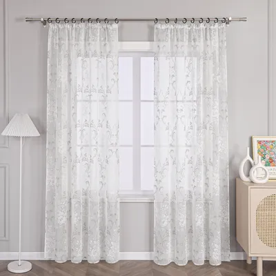 A pair of sheer curtains hanging in front of a window, with a floor lamp on the left and a cabinet with decor on the right.