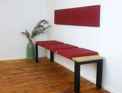 A modern bench with red cushions and a wooden seat is placed against a white wall, next to a green vase with dried branches.